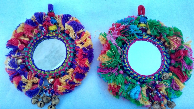 May include: Two round mirrors with colorful yarn tassels and beads. The mirrors are surrounded by a woven frame with a variety of colors, including red, blue, green, yellow, and purple. The mirrors are hanging from a string with a tassel.