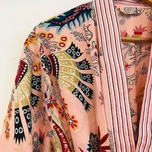 May include: A light pink kimono robe featuring a multi-colored floral and abstract pattern in shades of red, black, and olive green. The garment has a wide collar with vertical pink and white stripes and is displayed on a wooden hanger.
