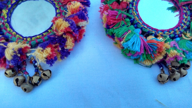 May include: Two round mirrors with colorful yarn fringe and small bells hanging from the bottom. The mirrors are surrounded by a woven design with a variety of colors, including purple, pink, yellow, green, and blue.