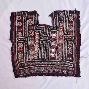 May include: A dark maroon textile with detailed embroidery and mirror embellishments. The design incorporates geometric patterns, circles, and rectangular shapes, with white, red, and blue accents. The edges are finished with a red border.