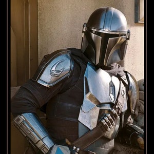 May include: A silver and black Mandalorian helmet with a visor. The helmet is worn by a person in a black and silver suit of armor. The armor has a symbol on the shoulder.