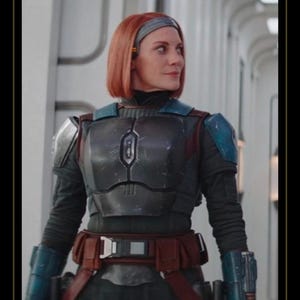 May include: A person with shoulder-length red hair wearing dark gray chest armor with blue shoulder plates, blue gauntlets, and a brown utility belt. The armor features a central vertical emblem on the chest plate and a dark long-sleeved undersuit.