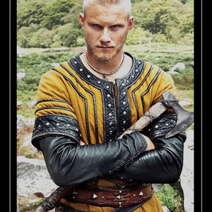 May include: A man with a shaved head and a beard is wearing a yellow and black leather tunic with a brown leather belt. He is holding a small axe in his right hand. The background is a blurry green forest.