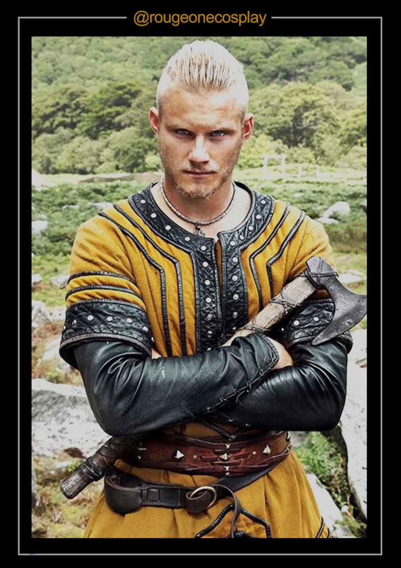 May include: A man with a shaved head and a beard is wearing a yellow and black leather tunic with a brown leather belt. He is holding a small axe in his right hand. The background is a blurry green forest.