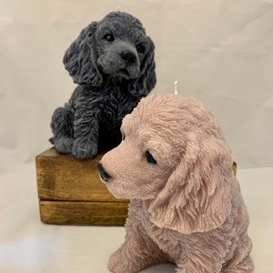 May include: Two adorable puppy candles, one in a dark grey colour and the other in a light pink colour. Both candles are sitting on a wooden box.