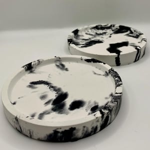 May include: Two circular coasters, white with a black marbled ink pattern. Each coaster has a raised edge and a smooth surface. The ink design varies, giving each coaster a unique appearance. These coasters are suitable for protecting surfaces from drink marks.