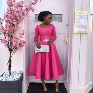 May include: A woman wearing a pink dress with a silver belt and a silver clutch. She is standing in front of a pink door with a sign that says "blossom".