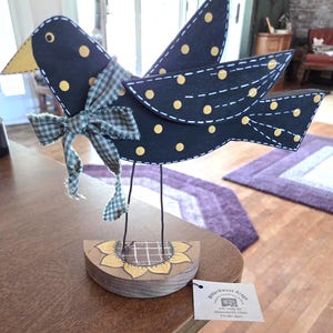 May include: A decorative black bird figurine with yellow polka dots and a green plaid bow. The bird has a yellow beak and is perched on a wooden base with a sunflower design. The bird's wings are outstretched, and it has wire legs.