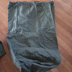 May include: An olive green canvas duffel bag with a drawstring closure and a metal ring. The bag is made of a durable, textured fabric and is designed to hold a large amount of items. The bag is shown on a wooden floor.