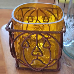May include: A yellow glass candle holder with a square shape and a rusty metal cage. The cage has a jack-o'-lantern face design. The candle holder is sitting on a wooden surface.