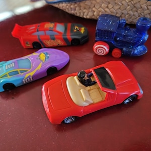 Puede incluir: Una colección de cuatro coches de juguete, que incluyen un coche de carreras rojo, un coche de carreras morado y azul con el número 101 en el lateral, un descapotable rojo con una figura de una persona en el asiento del conductor y un tren de juguete azul con una rueda de caramelo rojo y blanco.