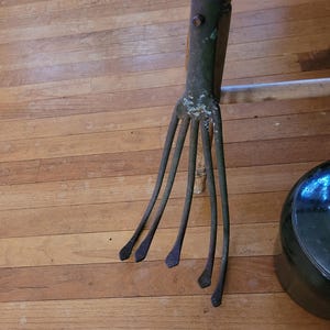 Könnte beinhalten: Ein rostiges Gartenwerkzeug aus Metall mit fünf Zinken. Das Werkzeug ist grün und braun mit einem Holzstiel.