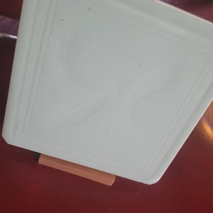 May include: A square, light-green glass tray with a raised border and a subtle embossed design. The tray is supported by a small, rectangular wooden stand, set against a red background. The tray's corners are slightly rounded.