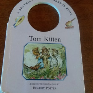 Könnte beinhalten: Ein rosa Türknaufbuch mit einer Beatrix Potter-Illustration von Tom Kitten. Der Buchtitel lautet "A Beatrix Potter Door Knob Book" und der Text "Tom Kitten" ist in einem schwarzen Kasten gedruckt. Das Buch enthält auch den Text "Based on the original tale by Beatrix Potter".