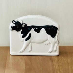 Op de afbeelding: Een witte keramische servetthouder in de vorm van een zwart-witte koe. De koe staat op alle vier de poten en heeft een zwarte neus en hoeven.
