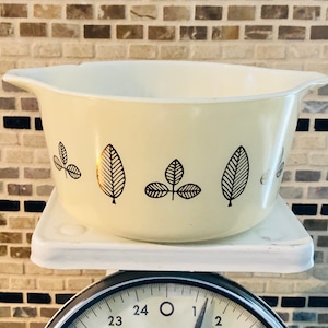 May include: Cream-colored vintage mixing bowl with black and gold leaf designs. The bowl has a flared rim and is sitting on a white square base. The bowl is a kitchenware item.