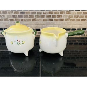 May include: A white ceramic sugar bowl with a yellow lid and a matching creamer. Both pieces have a yellow rim and green handles. Floral accents decorate the front of each piece. The sugar bowl has a lid with a small yellow knob.