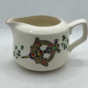 May include: White ceramic creamer with a gold rim and a Celtic knot design in yellow, red, blue, and green. The design includes shamrocks.