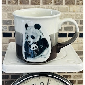 May include: A ceramic mug featuring a panda bear and cub illustration. The mug has a white interior and a handle with a gradient of brown and gray. The panda illustration is in black and white with a signature at the bottom.