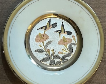 Könnte beinhalten: Ein kleiner, runder dekorativer Teller mit weißem Rand und goldenem Zierrat. Die Mitte zeigt einen silbernen Hintergrund mit einem Blumen- und Vogeldesign in Rosa-, Schwarz- und Goldtönen. Der Teller ist wahrscheinlich aus Porzellan.