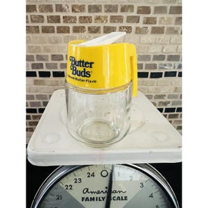 May include: A clear glass jar with a yellow lid and handle, labelled "Butter Buds Natural Butter Flavo." The jar is on a white surface, with a black and silver American Family Scale in the foreground.