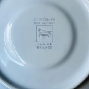 May include: A white porcelain saucer with a blue Longchamp logo. The logo features two horses running and the text "Longchamp Decor Inalterable Facon Main France".