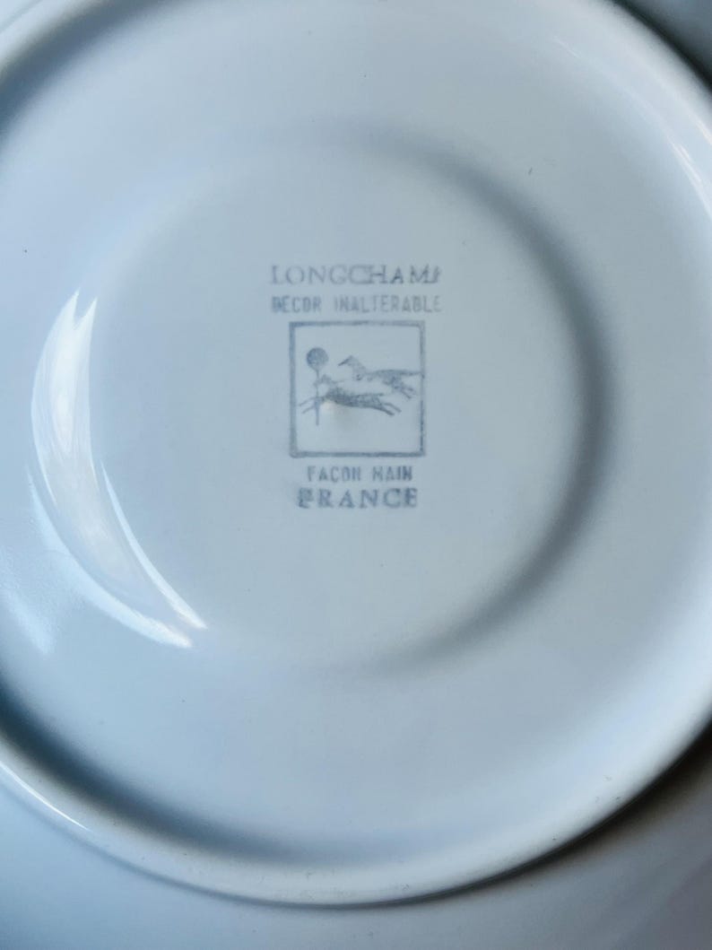 May include: A white porcelain saucer with a blue Longchamp logo. The logo features two horses running and the text "Longchamp Decor Inalterable Facon Main France".