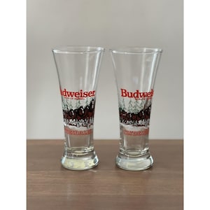 Puede incluir: Dos vasos de cerveza Budweiser transparentes. Cada vaso presenta el logotipo de Budweiser en rojo y blanco, con una ilustración de un carruaje tirado por caballos Clydesdale en marrón y blanco. Los vasos están sobre una superficie de madera.