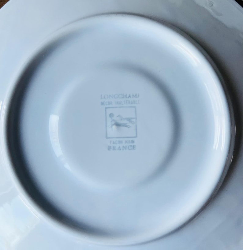 May include: White porcelain saucer with a Longchamp logo and the words "Decors Inalterables" and "Fait en France".