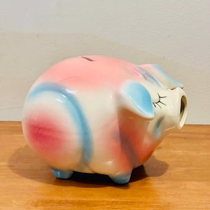 Pode incluir: Um cofrinho de cerâmica rosa e branco com detalhes azuis. O cofrinho tem uma fenda no topo para moedas e uma grande abertura lateral para recuperar as moedas.