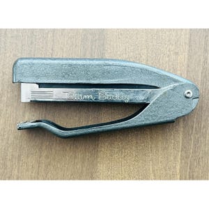 May include: A vintage, teal-colored stapler with the text "Tatum Buddy" printed on the side. The stapler is made of metal and has a classic design, with a lever and a staple ejection mechanism. The stapler is shown on a wooden surface.