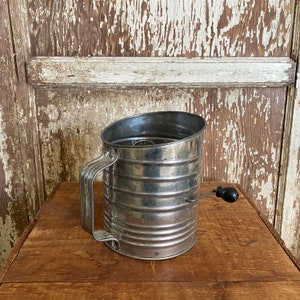 May include: A vintage silver-colored flour sifter with a black handle. The sifter has a round shape and a handle that extends to the side. The sifter is sitting on a wooden surface.
