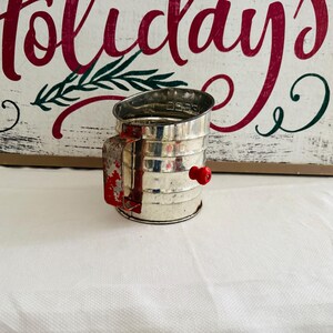 May include: A vintage silver metal flour sifter with a red handle and a red knob. The sifter has a mesh screen and a series of ridges around the side.