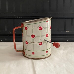 May include: Vintage white metal flour sifter with a red handle and a red knob. The sifter has a green and white checkered pattern with red flowers.