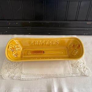 May include: Mustard yellow ceramic cracker serving dish with the word "CRACKERS" embossed on the rim. The dish has a built-in knife and cracker designs on each end. The dish is rectangular and sits on a white lace doily.