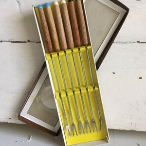 May include: A set of six fondue forks in a box. The forks have wooden handles with different colored tips: blue, white, and black. The box has a yellow interior and a brown exterior.