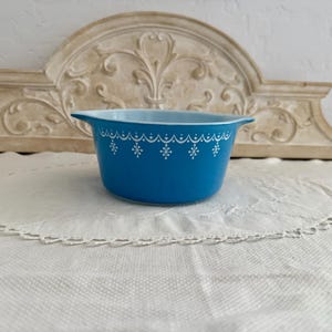May include: A vintage turquoise Pyrex bowl with a white snowflake and scalloped design. The bowl has a light blue interior and two handles. This retro kitchenware is perfect for serving or storage.