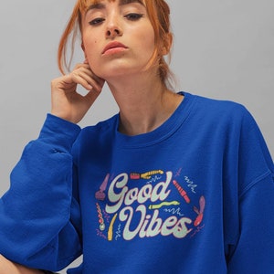 May include: A royal blue sweatshirt with the text "Good Vibes" in a retro font with colorful illustrations of flowers and other designs.