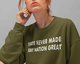 Pode incluir: Uma mulher vestindo uma camiseta verde com o texto "Hate Never Made Any Nation Great" em letras brancas. Ela está sentada em uma superfície cinza usando uma saia preta.