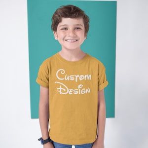 May include: A young person wearing a mustard yellow t-shirt with the text "Custom Design" in white. The person is standing in front of a teal background.