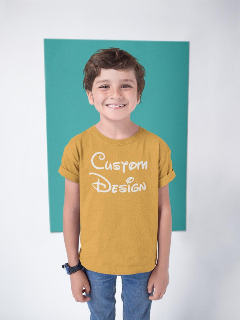May include: A young person wearing a mustard yellow t-shirt with the text "Custom Design" in white. The person is standing in front of a teal background.