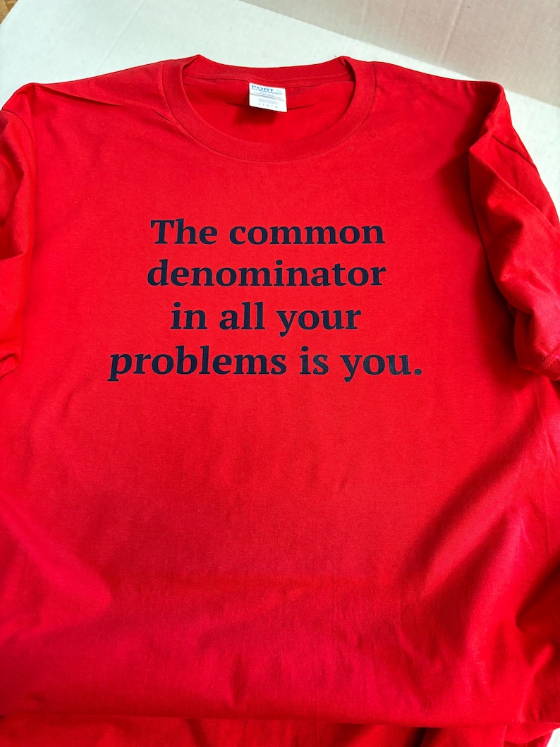 May include: Red t-shirt with black text that reads "The common denominator in all your problems is you."