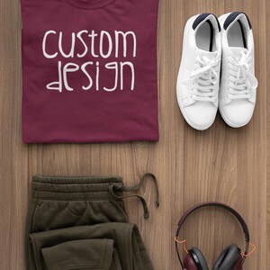 May include: A flat lay of a maroon t-shirt with the text "custom design" in white, a pair of olive green sweatpants, a pair of white sneakers, and a pair of burgundy headphones with black earcups.
