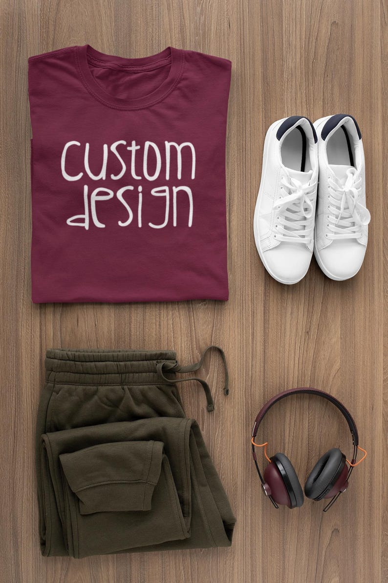May include: A flat lay of a maroon t-shirt with the text "custom design" in white, a pair of olive green sweatpants, a pair of white sneakers, and a pair of burgundy headphones with black earcups.