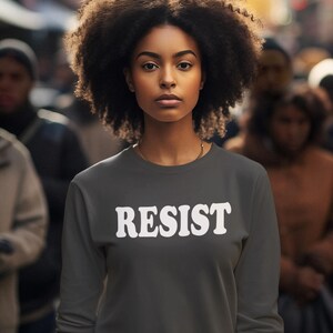Pode incluir: Uma mulher vestindo uma camiseta cinza escura com a palavra "RESIST" impressa em branco na frente. Ela tem cabelo escuro e encaracolado e est&aacute; em p&eacute; em uma multid&atilde;o de pessoas.
