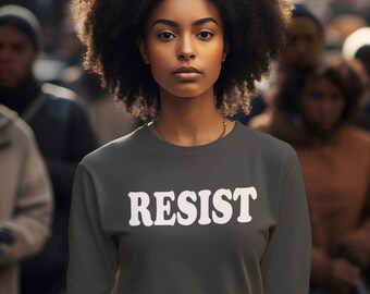 Pode incluir: Uma mulher vestindo uma camiseta cinza escura com a palavra "RESIST" impressa em branco na frente. Ela tem cabelo escuro e encaracolado e está em pé em uma multidão de pessoas.