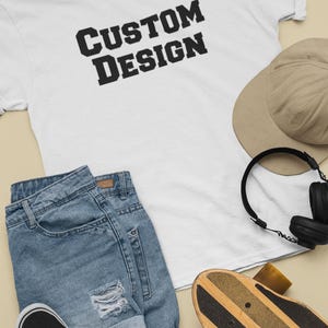 May include: A white t-shirt with the text "CUSTOM DESIGN" printed in black. The t-shirt is laid out on a beige surface with a pair of blue denim shorts, a brown baseball cap, black headphones, and a wooden skateboard.
