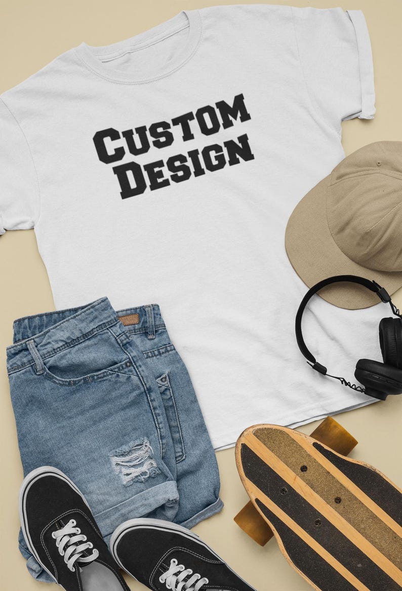 May include: A white t-shirt with the text "CUSTOM DESIGN" printed in black. The t-shirt is laid out on a beige surface with a pair of blue denim shorts, a brown baseball cap, black headphones, and a wooden skateboard.