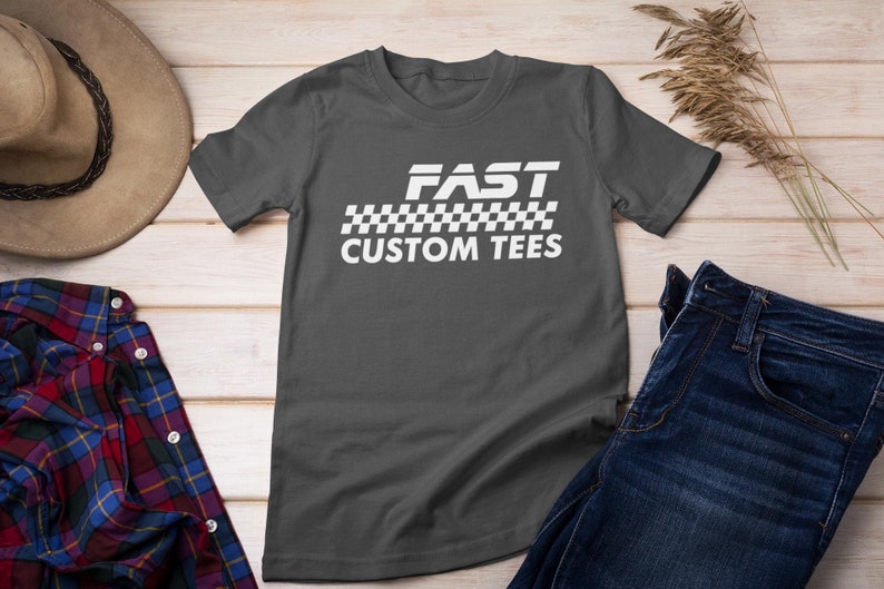 May include: A charcoal gray t-shirt with a white graphic that says "FAST" with a checkered flag design and the words "CUSTOM TEES" underneath.
