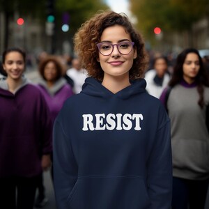 Pode incluir: Uma camisola de capuz azul marinho com a palavra "RESIST" impressa em branco na frente. A modelo est&aacute; a usar a camisola e tem cabelo castanho encaracolado e est&aacute; a usar &oacute;culos.